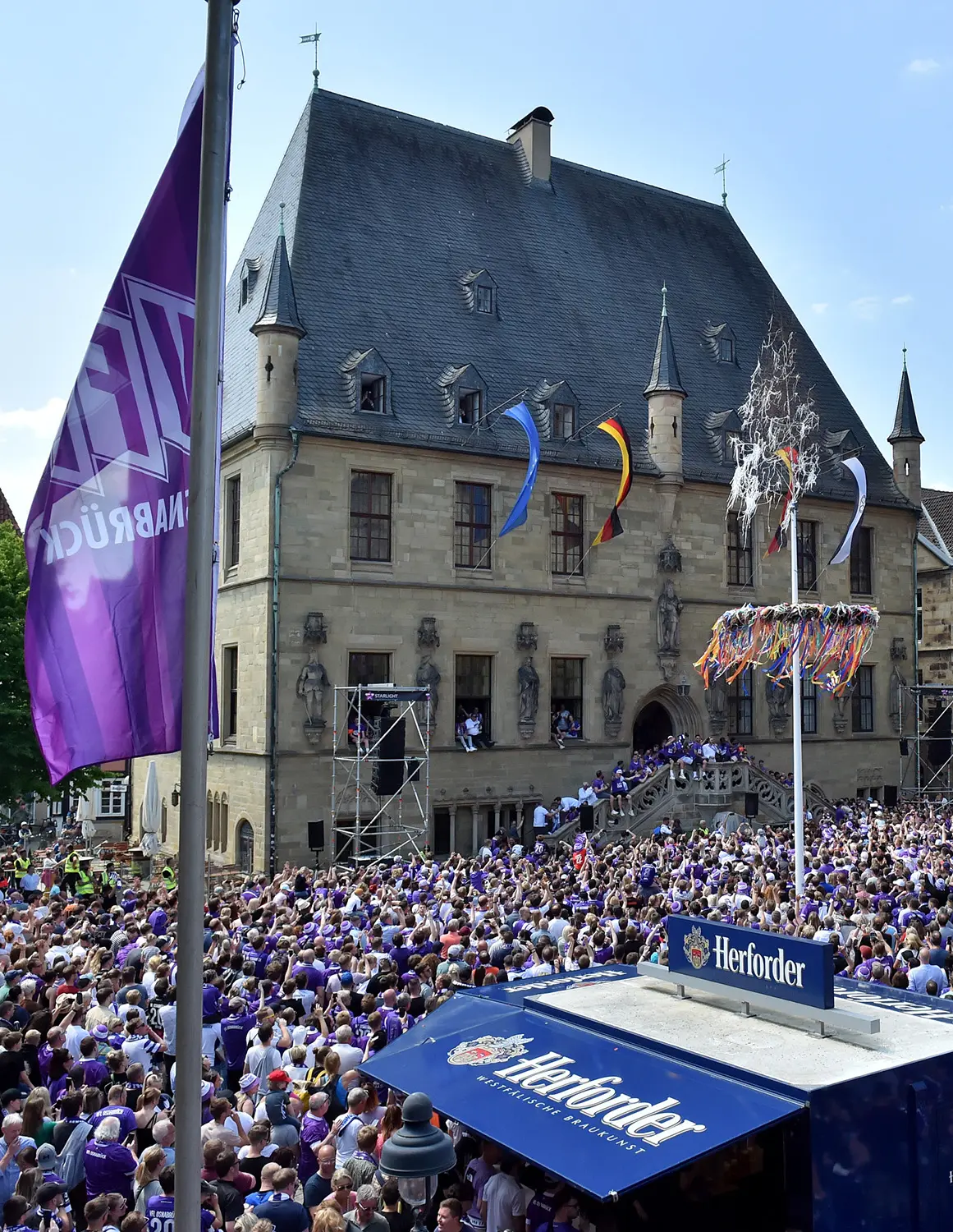 20230528_osnapix_59_hoch Große Menschenmenge vor historischem Gebäude, Fahne und Herforder-Stand im Vordergrund.