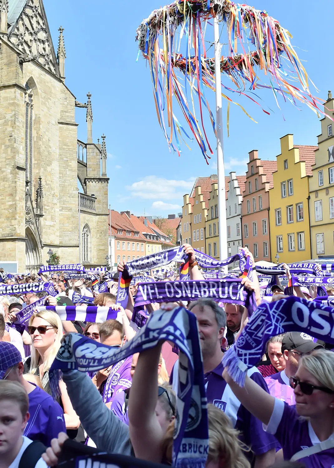 20230528_osnapix_62_hoch Menschenmenge mit VfL Osnabrück Schals vor Häusern und Kirche. Maibaum mit bunten Bändern im Hintergrund.