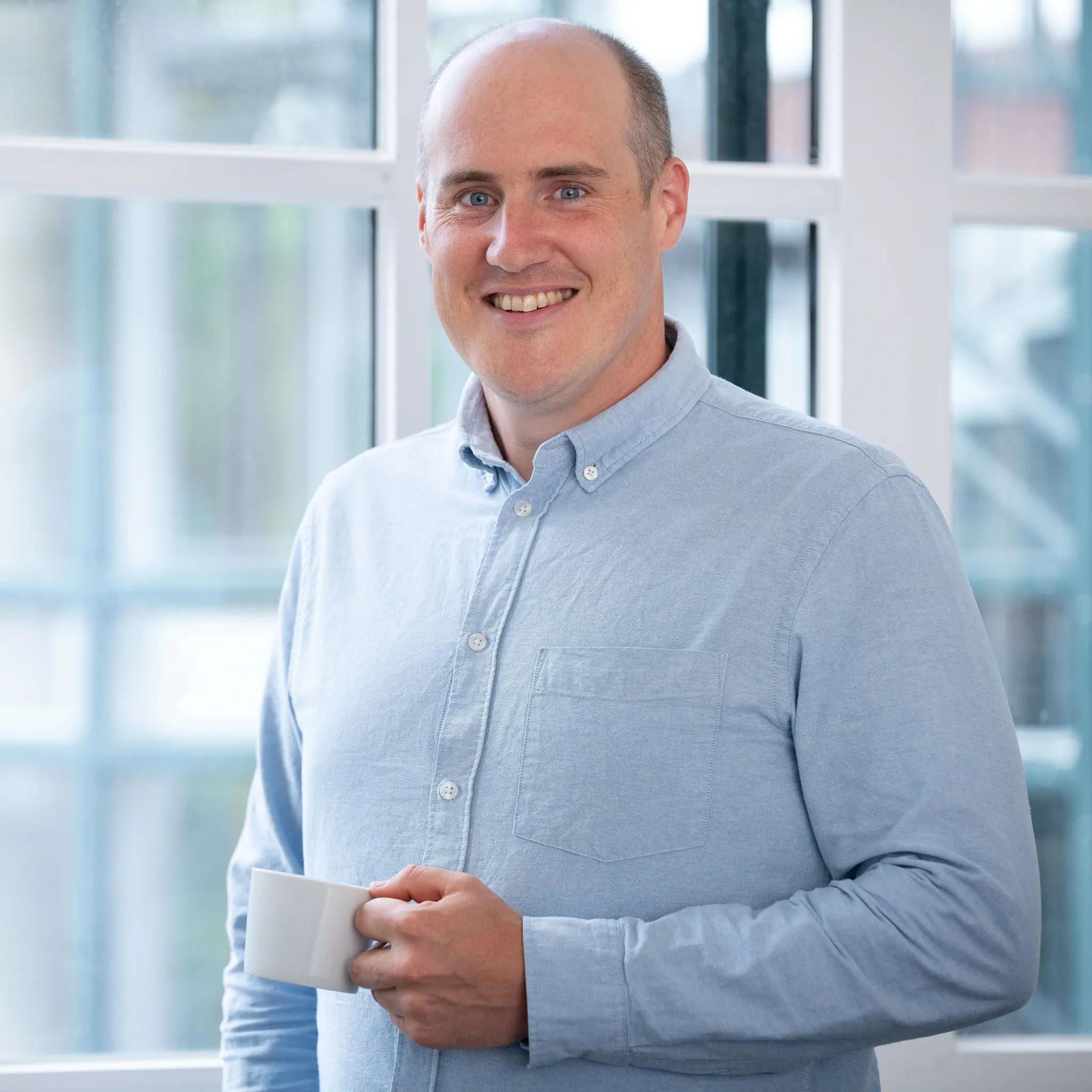 Tom Herter, Porträt. Mann lächelt mit Tasse in der Hand vor Fenster.